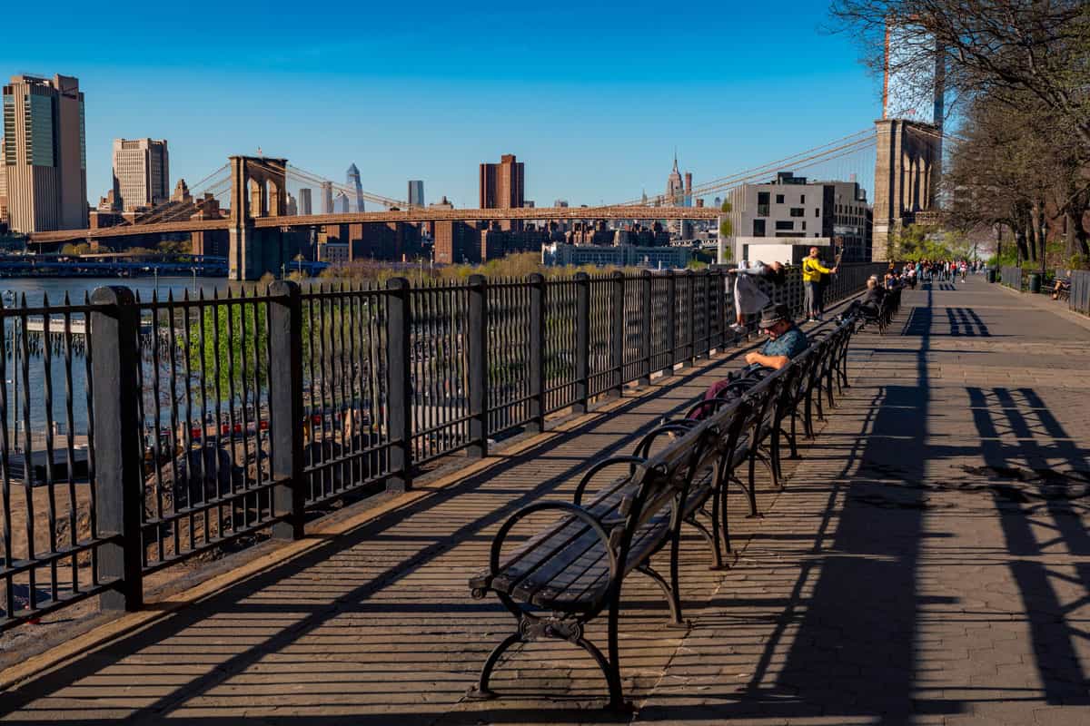 man sitting on long line of benches at sunset overlooking Brooklyn Bridge, Manhattan and East River in Brooklyn Heights neighborhood