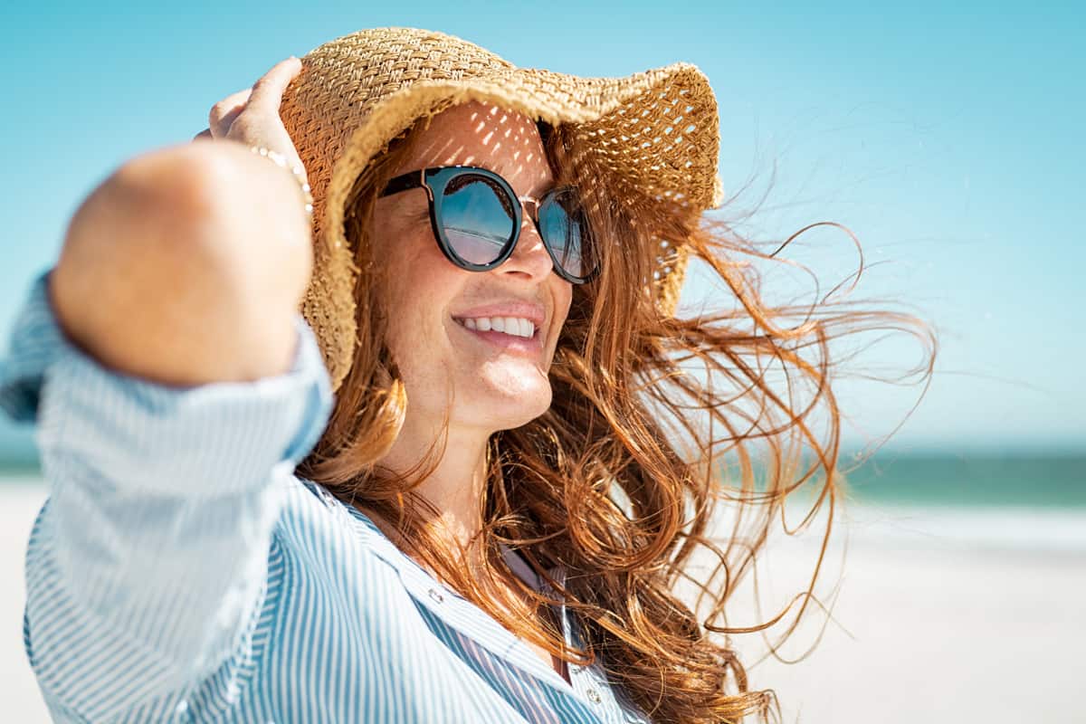 Side view of beautiful mature woman wearing sunglasses enjoying at beach. Young smiling woman on vacation looking away while enjoying sea breeze wearing straw hat.