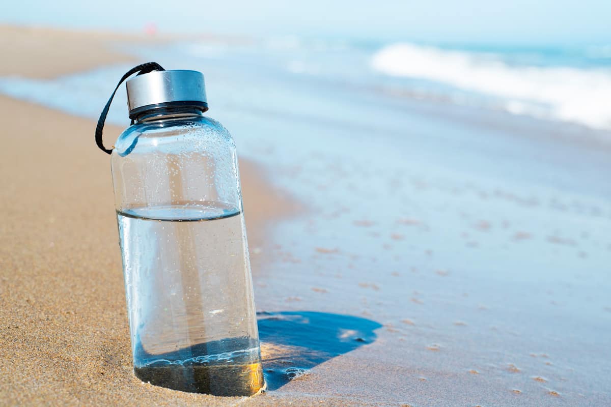closeup of a glass reusable water bottle on the seashore of a lonely beach