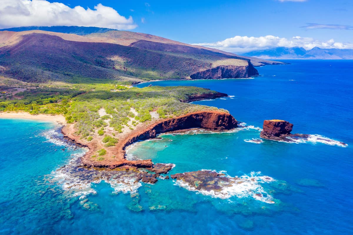 Aerial view of Lanai, Hawaii featuring Hulopo'e Bay and beach, Sweetheart Rock (Pu'u Pehe), Shark's Bay, and the mountains of Maui in the background. 