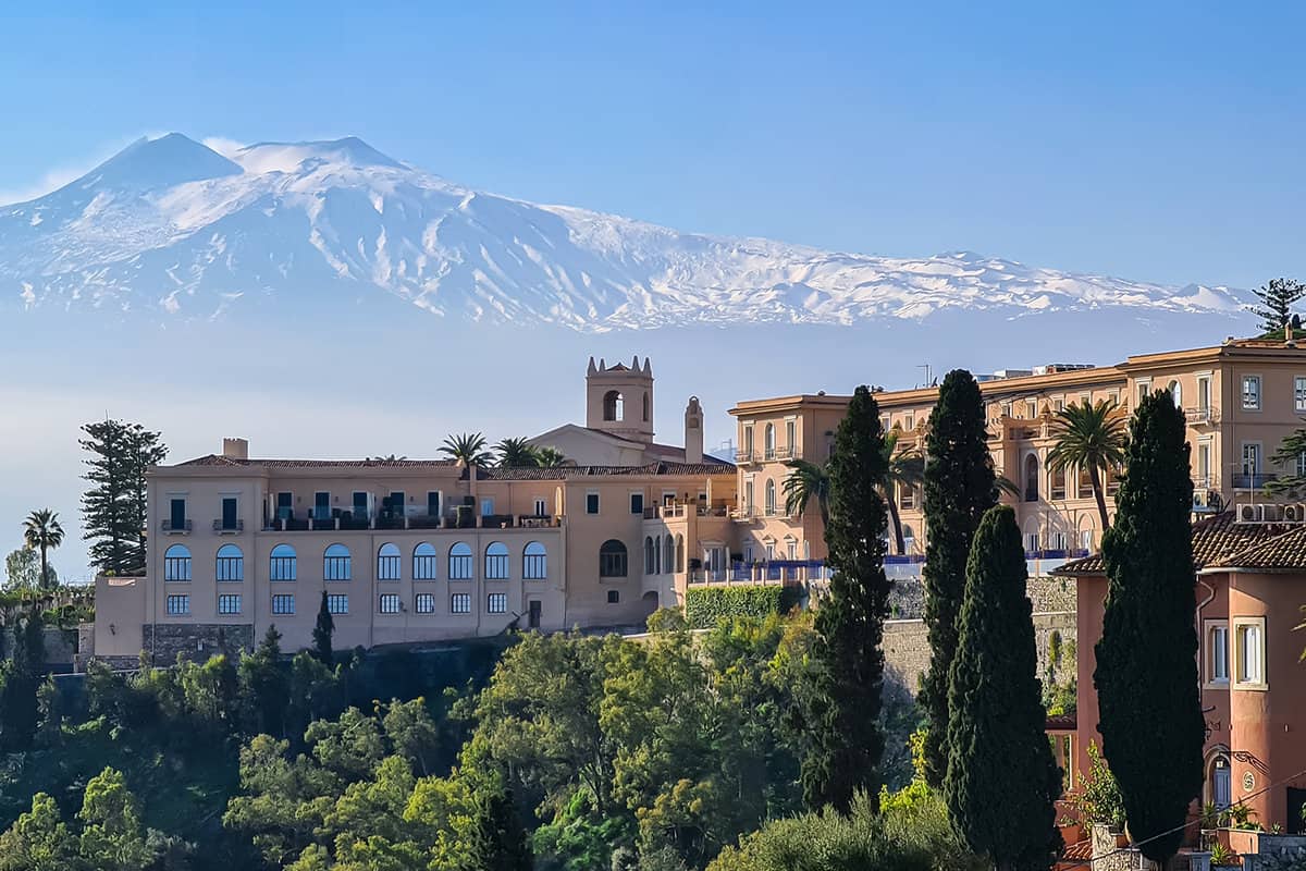 Luxury San Domenico Palace Hotel with panoramic view on snow capped Mount Etna volcano on sunny day from public garden Parco Duca di Cesaro to Giardini Naxos in Taormina, Sicily, Italy