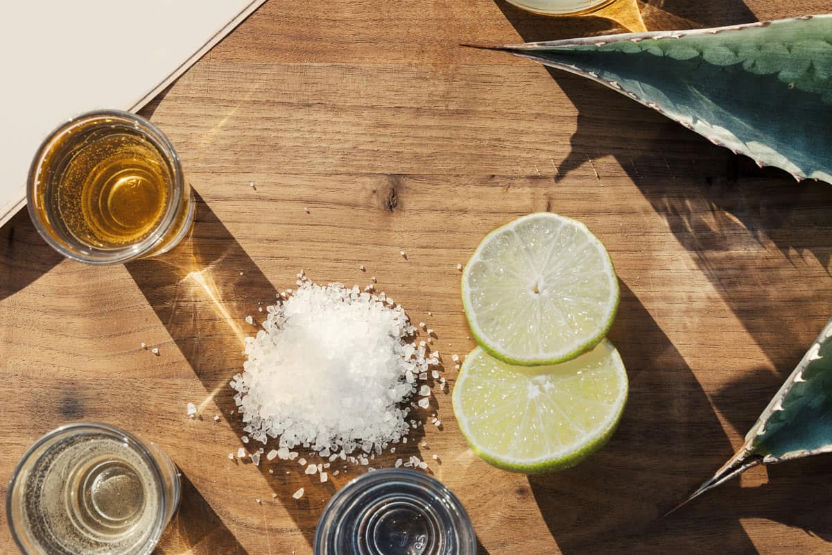 Overhead view of three kinds of tequila on an outside table with lime salt and agave leaves.