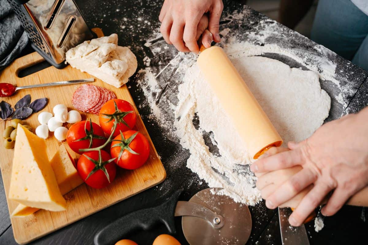 Roll out pizza dough, cooking pizza with a kitchen rolling pin on a black countertop at home. Ingredients for Italian pizza papperoni recipe.