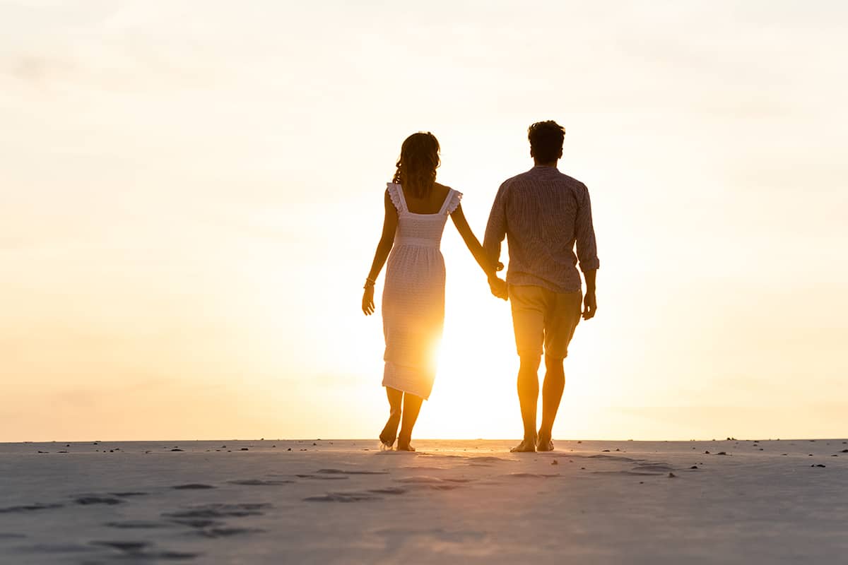 Silhouettes of man and woman holding hands while walking on beach against sun during sunset