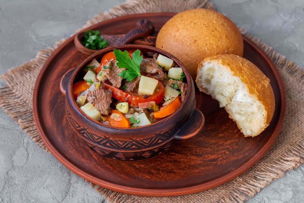 Vegetable beef stew with potato, carrots, tomatoes, onion and herbs served in a clay pot with white bread rolls. Selective focus, horizontal. 