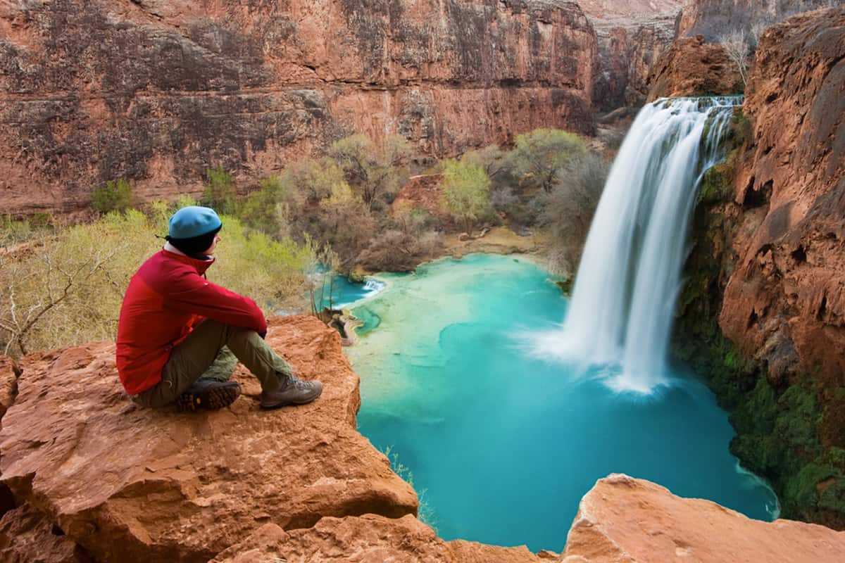 Woman sitting at the edge of a cliff watching Havasu Falls drop into it's turquoise pool. Havasu Canyon, Arizona, Mooney Falls in Havasu Indian reservation, Havasu Falls Reopens in Grand Canyon: Plan Your Visit Now!