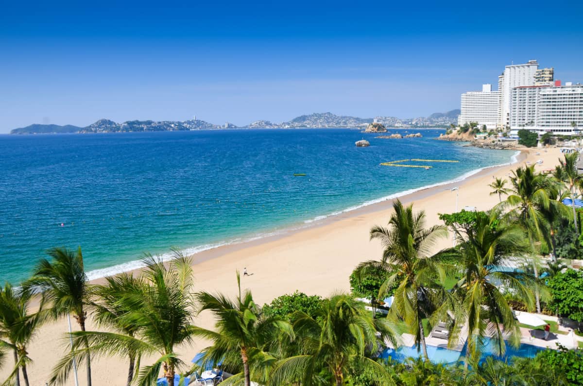 Beautiful view of the beach in Acapulco. Palm trees, crisp blue waters, and a hotel in the distance.