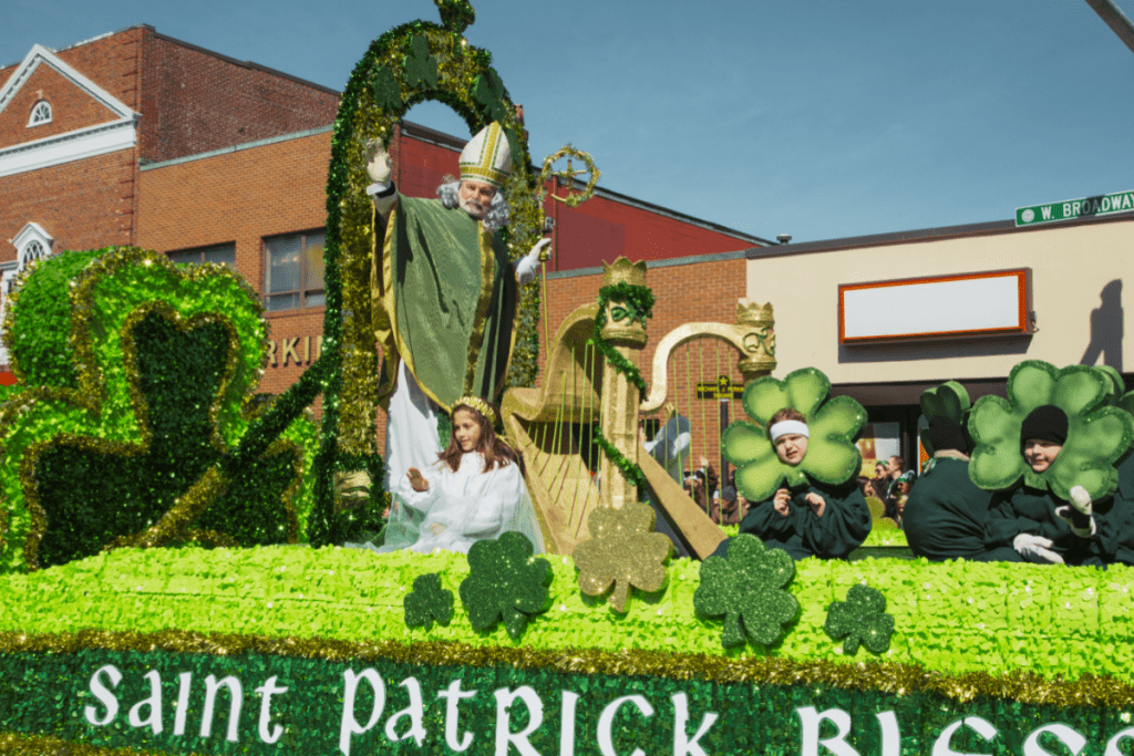 A parade float for St. Patrick's Day. It is covered in green clover. it has large shamrock decorations, and features someone dressed up as St. Patrick blessing a crowd in Boston, MA