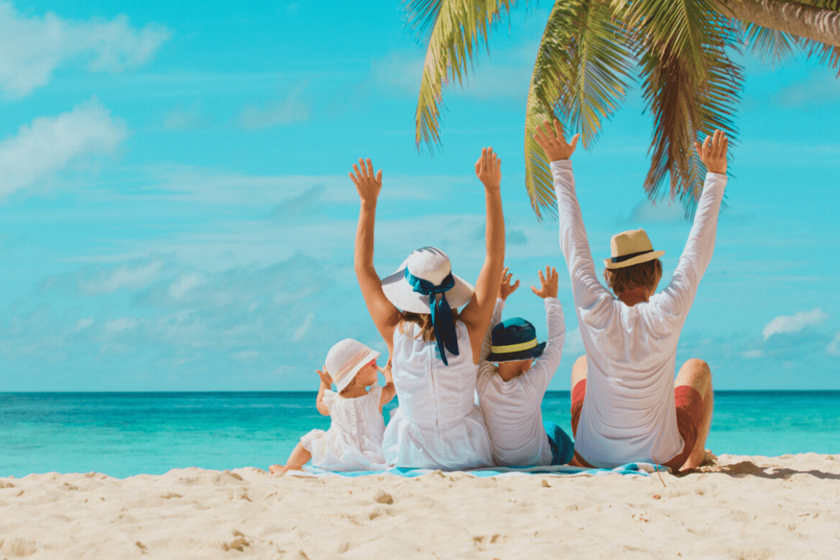 Summer travel photo of a family on the beach. There is a family of four on the beach looking out into the ocean. They are all wearing white and sun hats.
