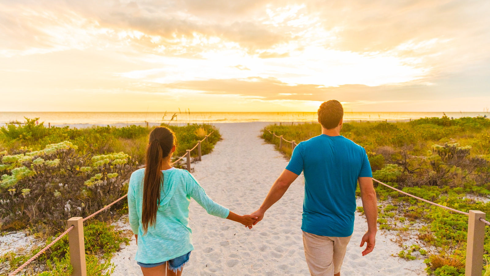 Happy young couple in love walking on romantic evening beach stroll at sunset. Lovers holding hands on summer holidays in Florida beach vacation destination. People walking from behind 1600x900