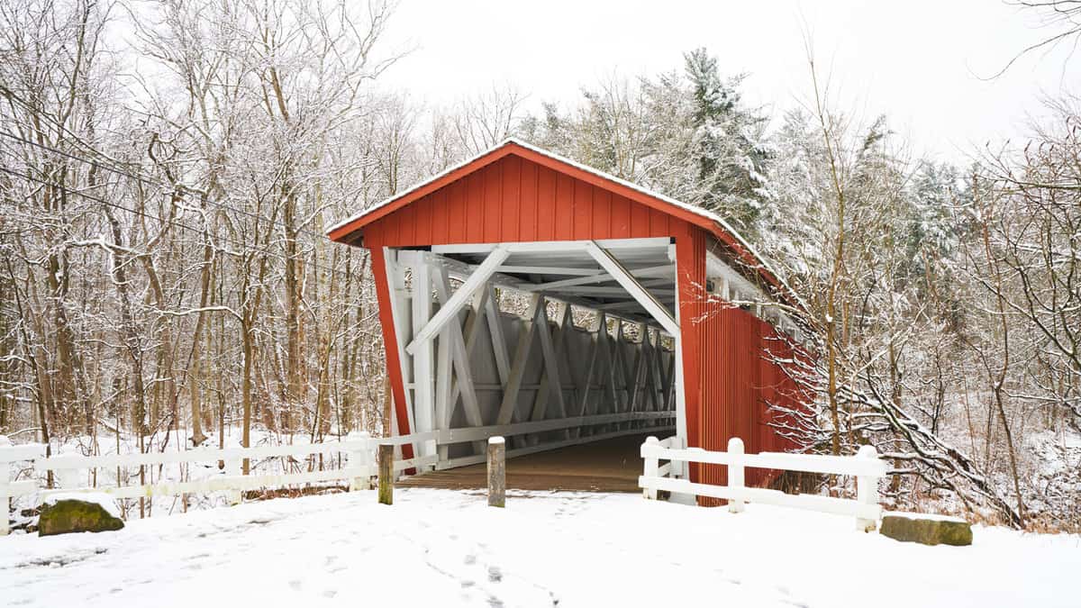 The everett road covered bridge in Cuyahoga Valley National Park. The bridge is red with a white interior. It is a snowy day, and the nearby trees in the forest are covered with fluffy white.