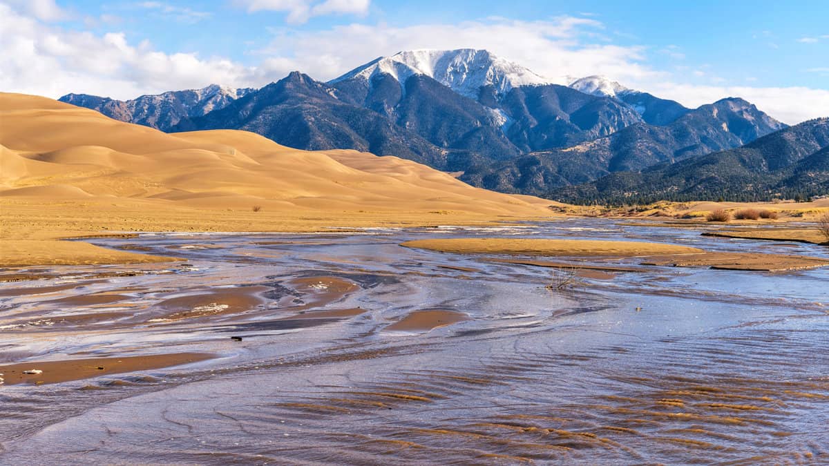 A bright sunny Spring morning view of Medano Creek rushing down a sandy valley at base of rolling Great Sand Dunes