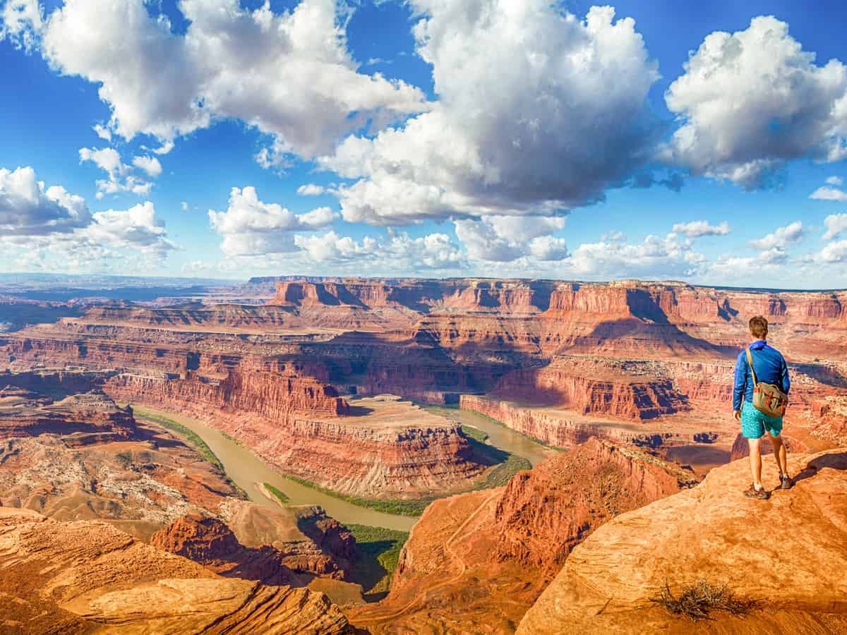 Panoramic view of young hiker standing on a cliff in scenic Dead Horse Point State Park enjoying the view on a beautiful sunny day with blue sky and dramatic clouds in summer, Utah, USA