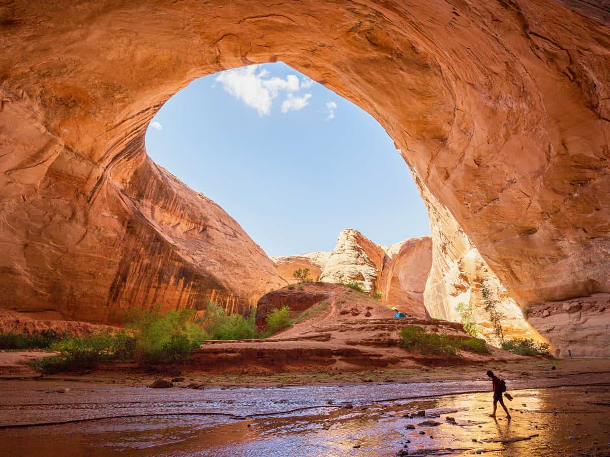Wide angle view of male hiker backpacking beneath stunning Jacob Hamblin Arch in Coyote Gulch on a sunny day with blue sky and clouds in summer, Grand Staircase-Escalante National Monument, Utah, USA