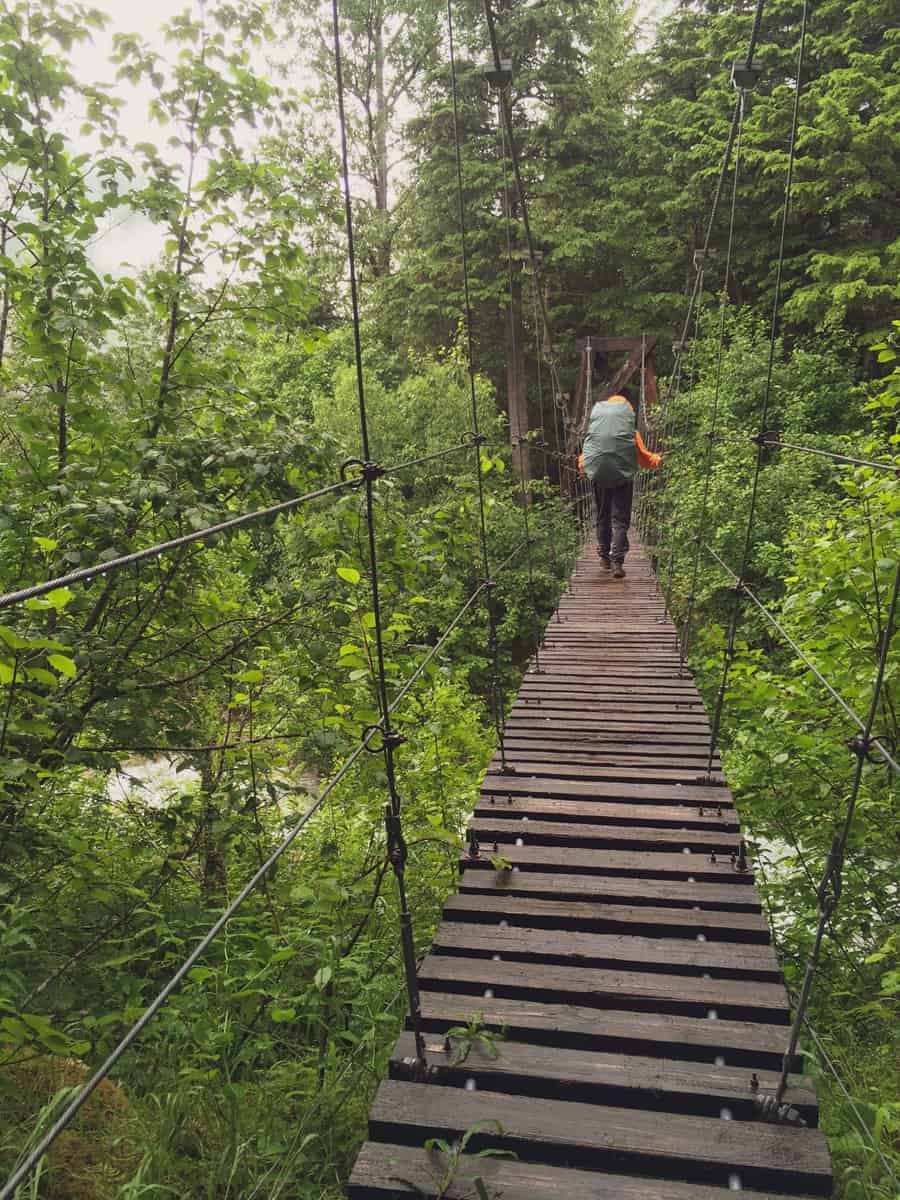 A teenage boy with a heavy backpack crosses a river on wobbly suspension bridge while backpacking the Klondike Gold Rush's historic chilkoot trail on a rainy summer holiday. Skagway, Alaska, USA.