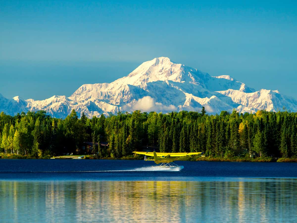 A yellow seaplane (float plane) landing on Kashwitna Lake with Mount McKinley or Denali in the background, Alaska