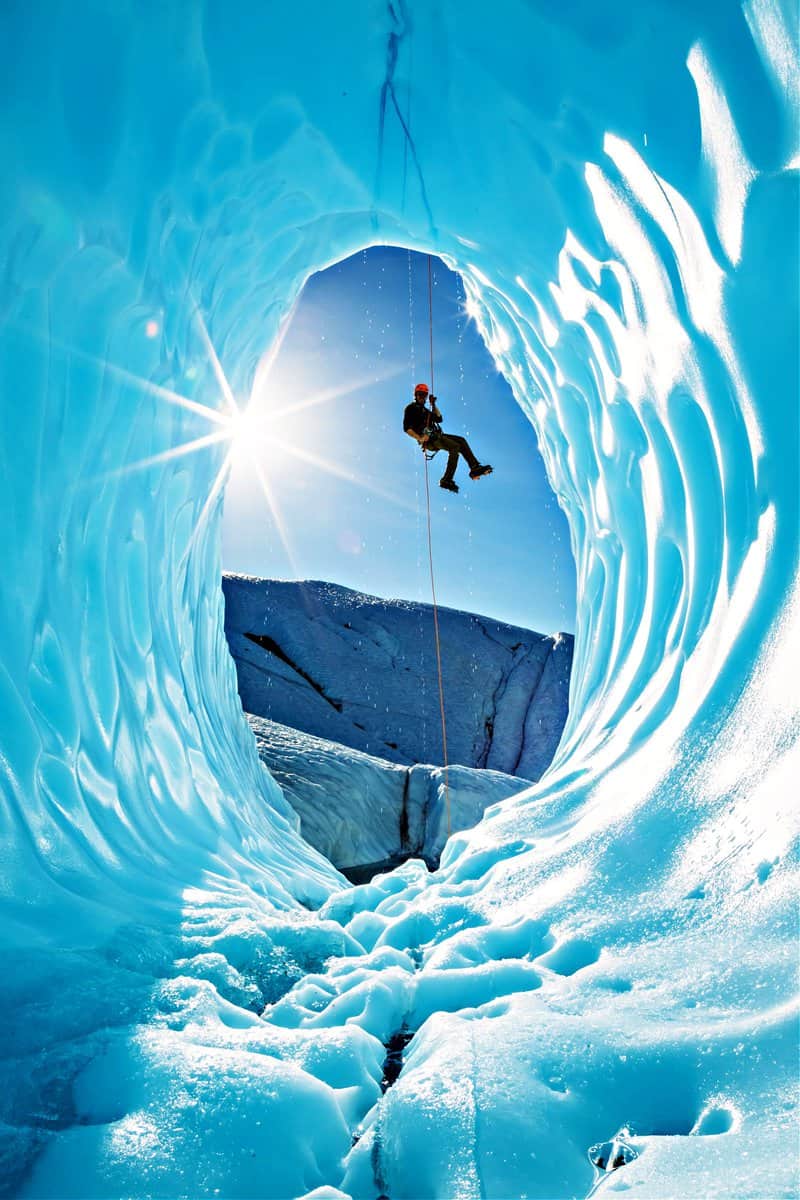 Active young man ice climber on glacier rappelling into ice cave entrance in Alaska.