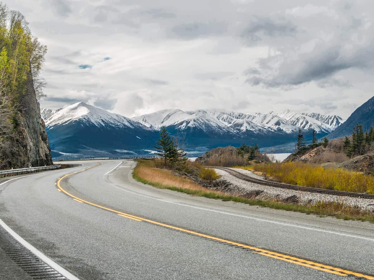 Alaska Scenic Road: The Seward Highway curves beneath cloudy skies as it passes by snow-covered mountains at the edge of an ocean inlet south of Anchorage.