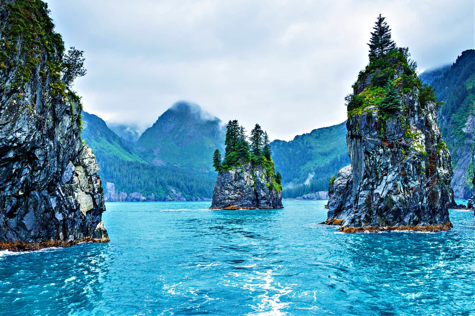 Blue waters and tree covered rocks jutting out of water on a cloudy morning at Porcupine bay at Kenai Fjords National Park, Alaska