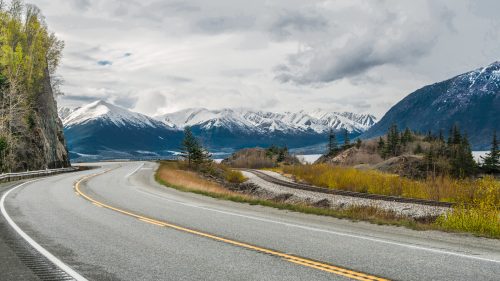 The Seward Highway curves beneath cloudy skies as it passes by snow-covered mountains at the edge of an ocean inlet south of Anchorage 1600x900