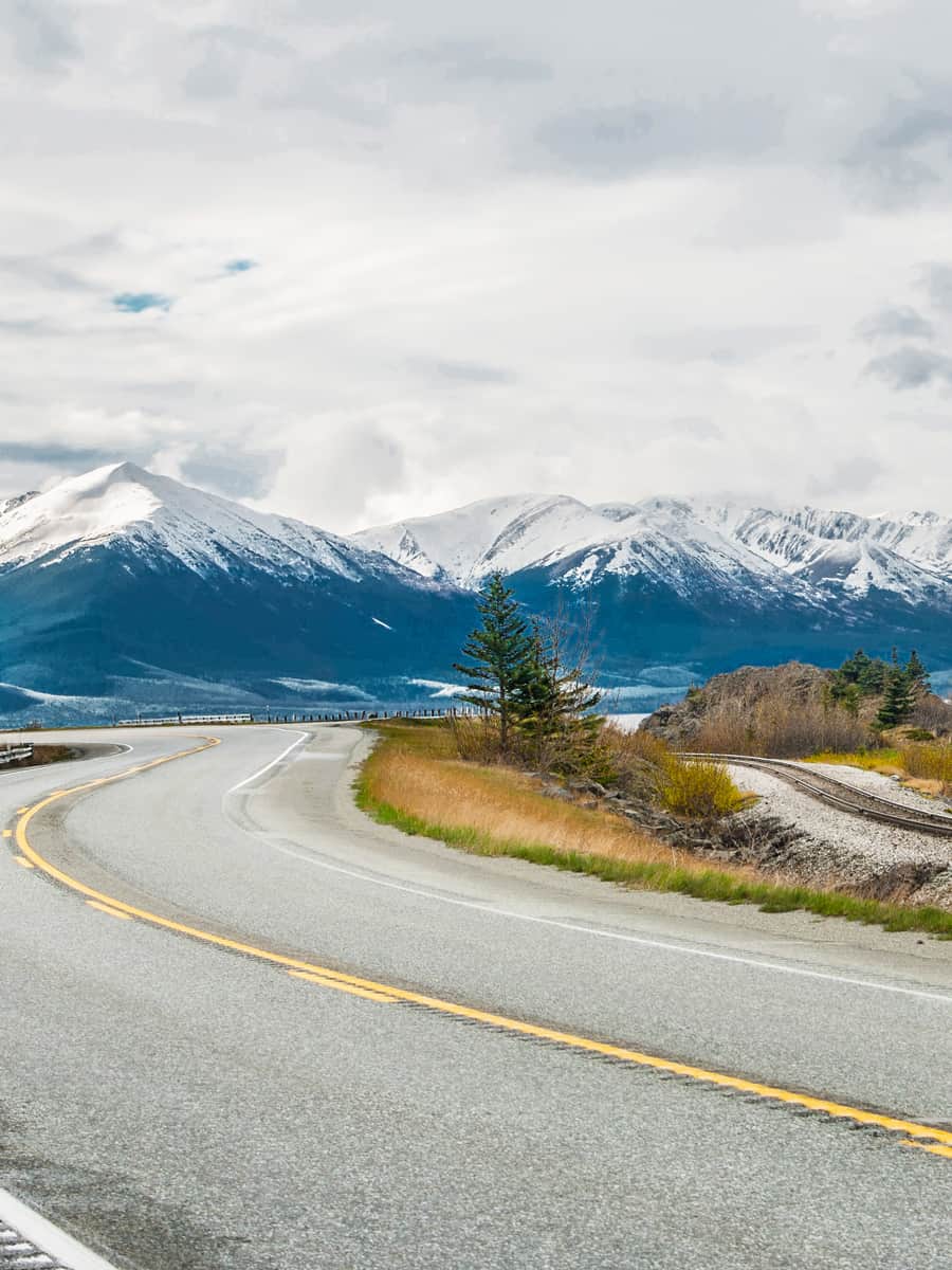 The Seward Highway curves beneath cloudy skies as it passes by snow-covered mountains at the edge of an ocean inlet south of Anchorage