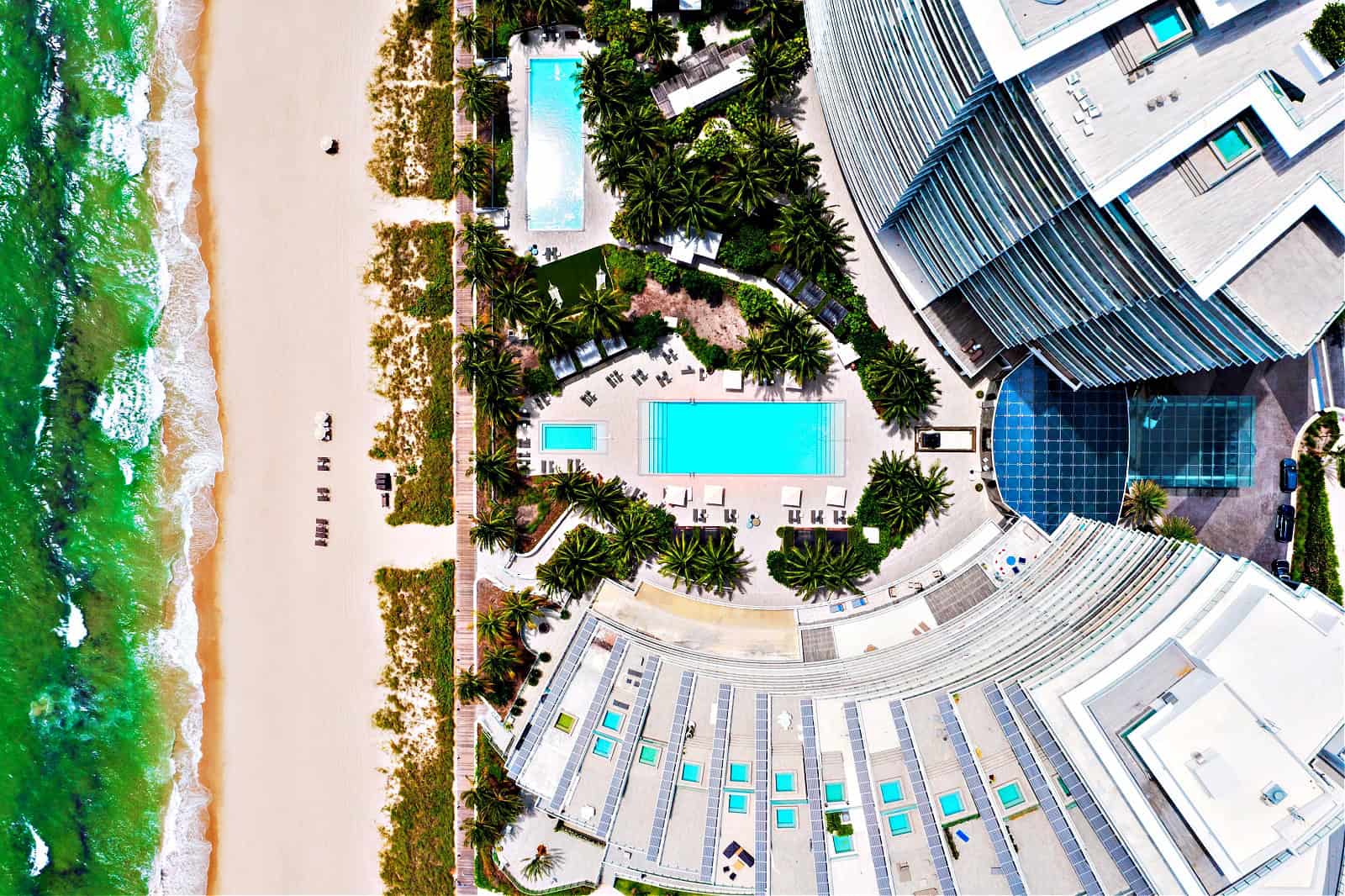 Aerial view of a beautiful pool area in a modern luxury building, palm trees, green areas, sun loungers, umbrellas, tropical plants, balconies, covered patio, paths to the beach, hammocks