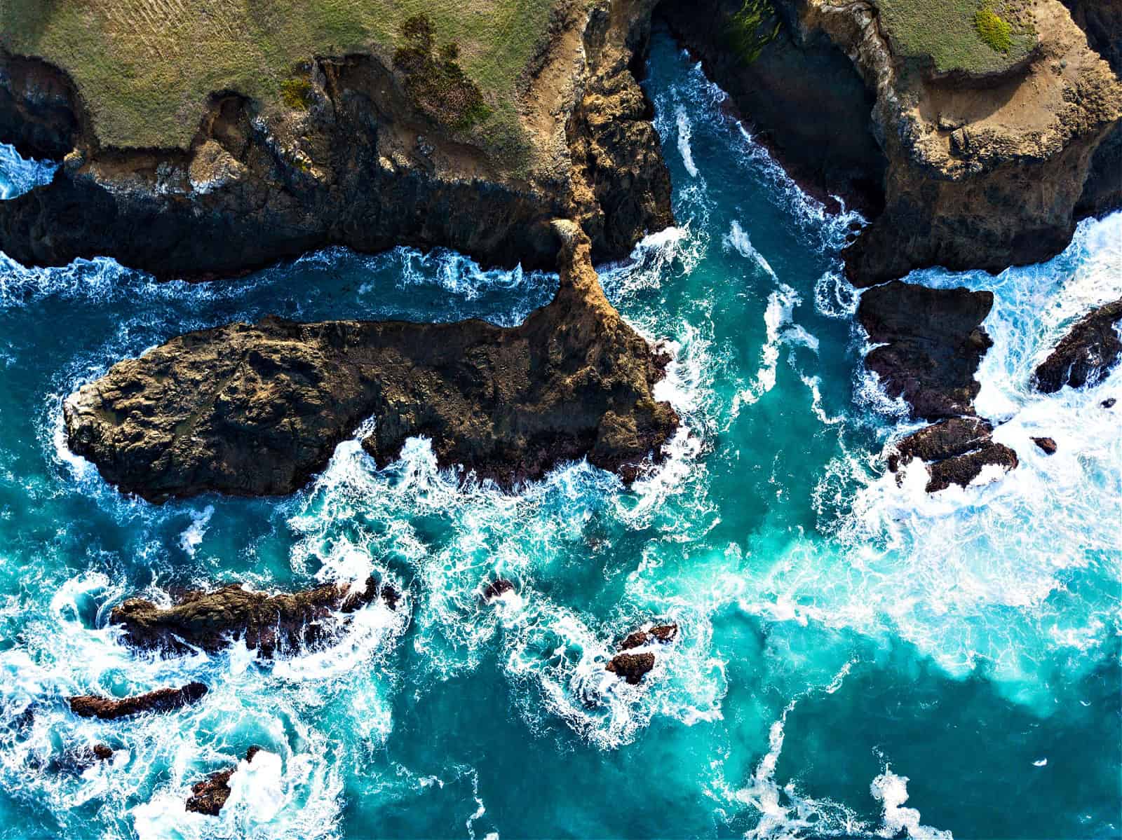 An aerial view of the Mendocino coast in northern California shows the cold, colorful waters of the Pacific Ocean crashing against the rocky shoreline.
